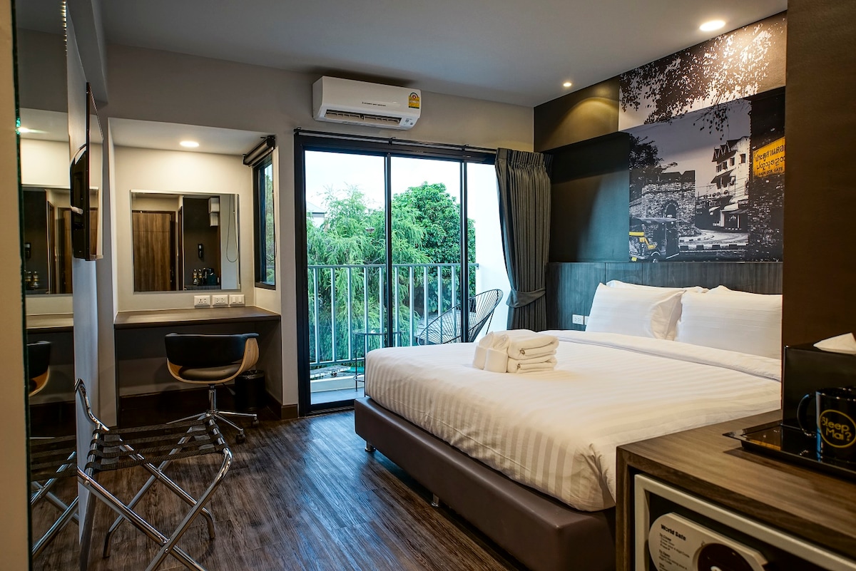 A modern balcony room features large windows that provide natural light, highlighting the Sealy Posturepedic mattress on the spacious bed. A flat-screen TV is mounted on the wall, and a mini fridge is conveniently placed. A seating area by the window offers views of greenery.