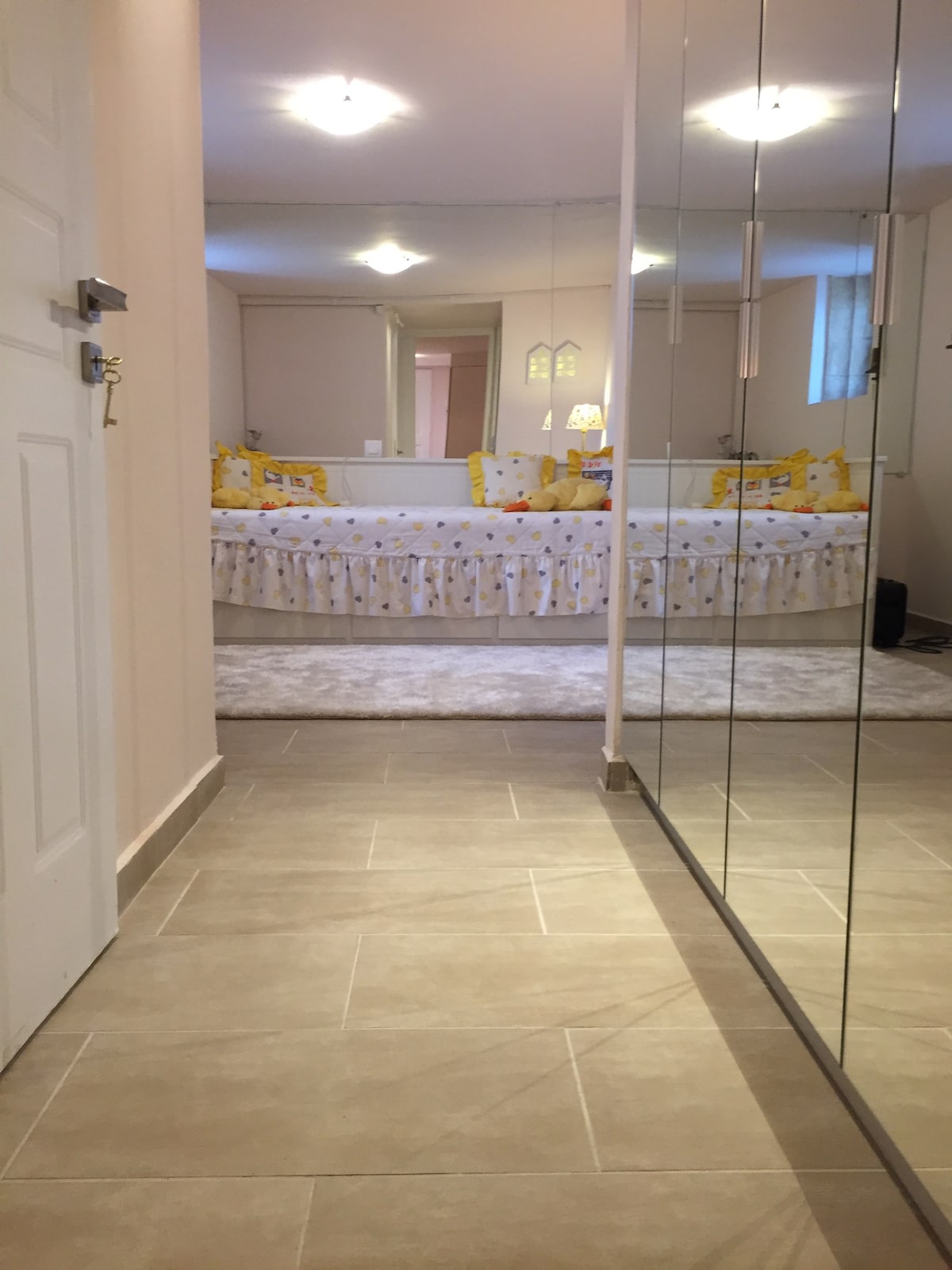 A well-lit entrance area leads to a room featuring a spacious bed adorned with yellow and patterned pillows. Mirrored panels enhance the sense of space, while neutral-toned tiles provide a clean and modern look. Soft lighting is provided by overhead fixtures.