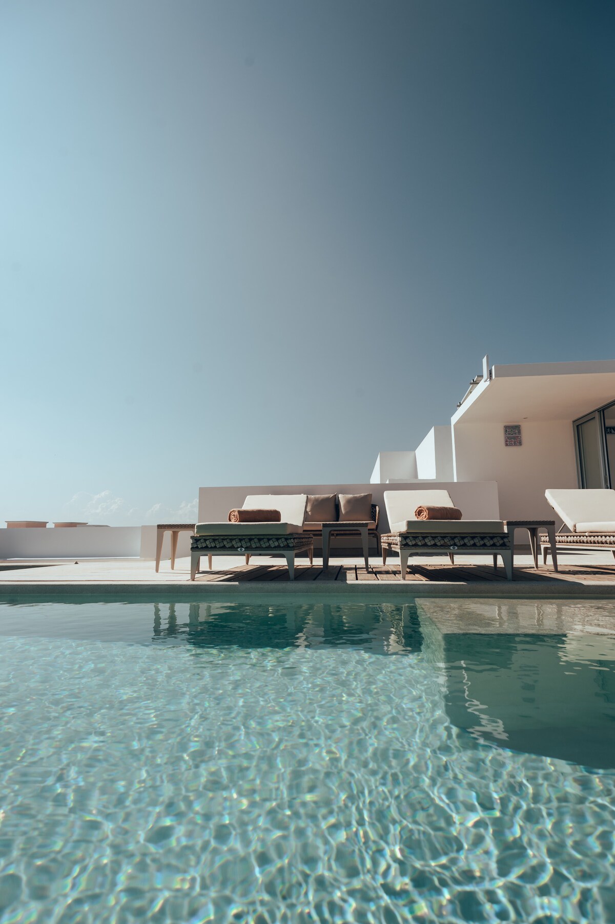 Stylish lounge chairs are positioned beside a serene pool, reflecting the clear blue sky. The inviting pool area creates opportunities for relaxation, with gentle waves lapping at the edge, providing a connection to the surrounding environment.