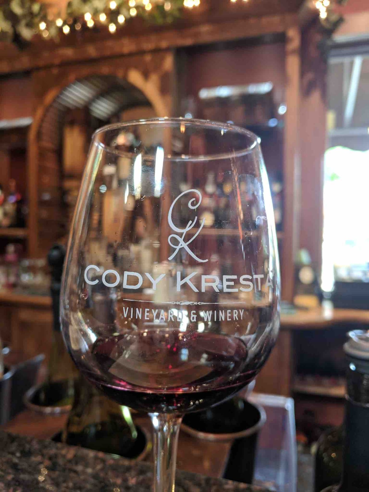 A wine glass displaying the CODY KREST VINEYARD & WINERY logo is filled with deep red wine. The background shows wooden shelves filled with bottles, creating a warm and inviting setting.