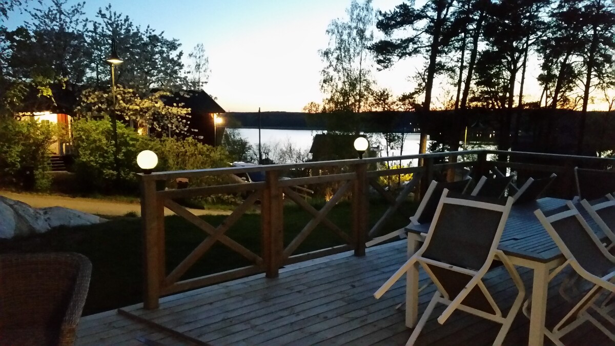 A wooden sun deck is featured in the foreground, furnished with white chairs and a table. Soft lighting glows from spherical lamps, and the view of the water is visible in the distance. The trees frame the scene, enhancing the serene environment at dusk.