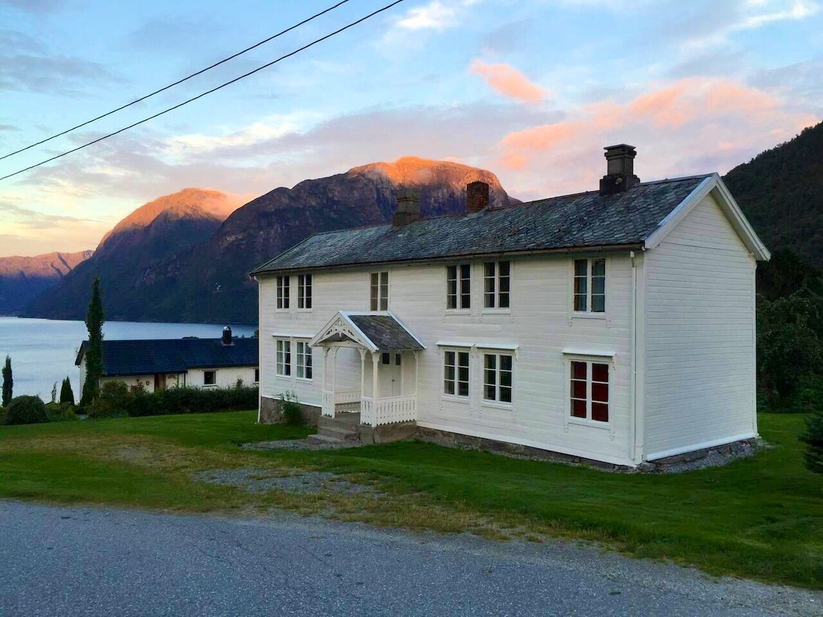 A white, two-story house features evenly spaced windows and a welcoming entryway with a porch. The surrounding lawn provides ample green space. In the background, mountains frame the horizon, complementing the serene view of the nearby water.
