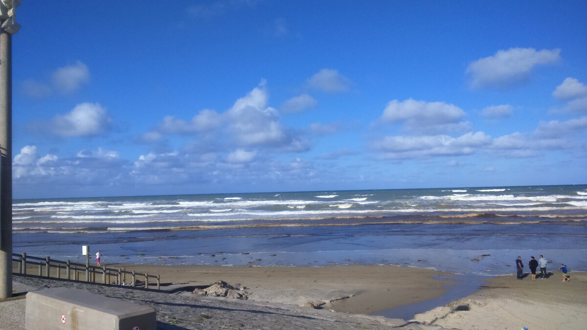 The beach scene features gentle waves lapping at the sandy shore under a bright blue sky with scattered clouds. A minimal number of people can be seen enjoying the coastline, with a promenade visible in the foreground leading towards the water.