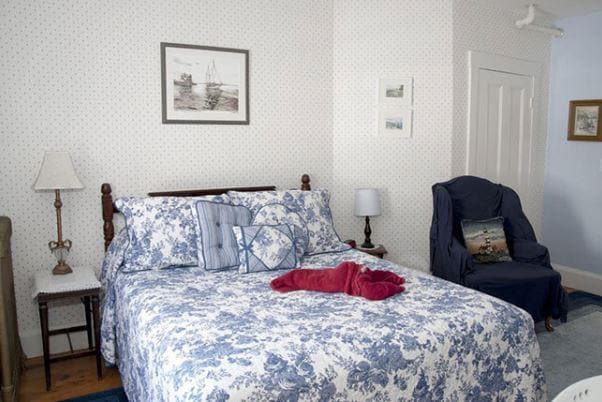 A spacious bed is arranged with a floral patterned coverlet and a red throw blanket. Accent pillows are neatly positioned. A bedside lamp and a chair complete the room's decor. Soft wall colors and framed nautical artwork add subtle charm.