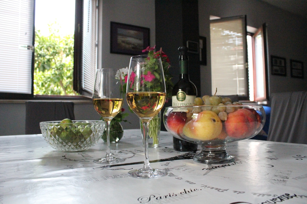 A dining table is set with two glasses of wine beside a bowl of assorted fresh fruit, including apples and grapes. The table is adorned with a decorative tablecloth. Sunlight filters through partially opened windows, illuminating the inviting space.