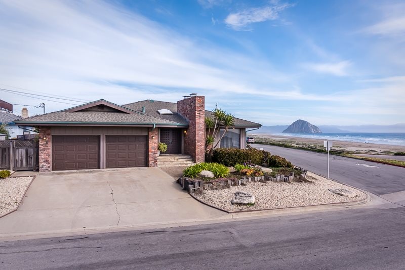 A single-level oceanfront beach house is visible, displaying a brick exterior with a neatly landscaped yard. The driveway leads to a two-car garage, flanked by rocks and plants. In the background, ocean views and Morro Rock are seen under a clear sky.