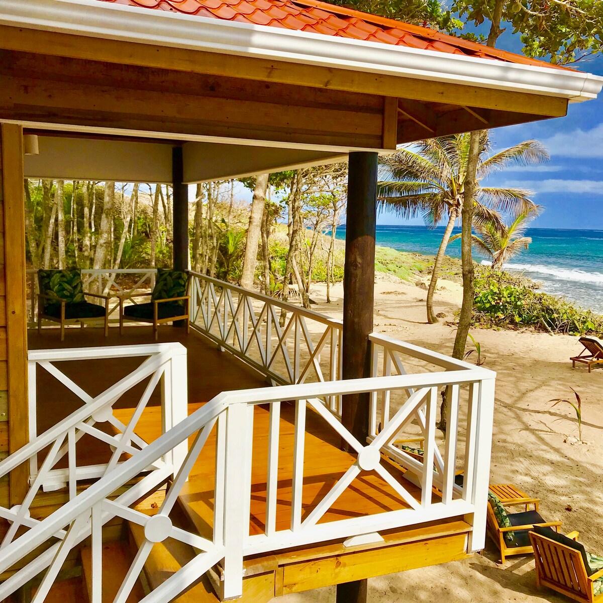 A wooden deck is featured with a white railing. The beach is visible just beyond, showcasing soft sand and waves gently lapping at the shore. Lush greenery and palm trees surround the area, creating a natural seaside setting.
