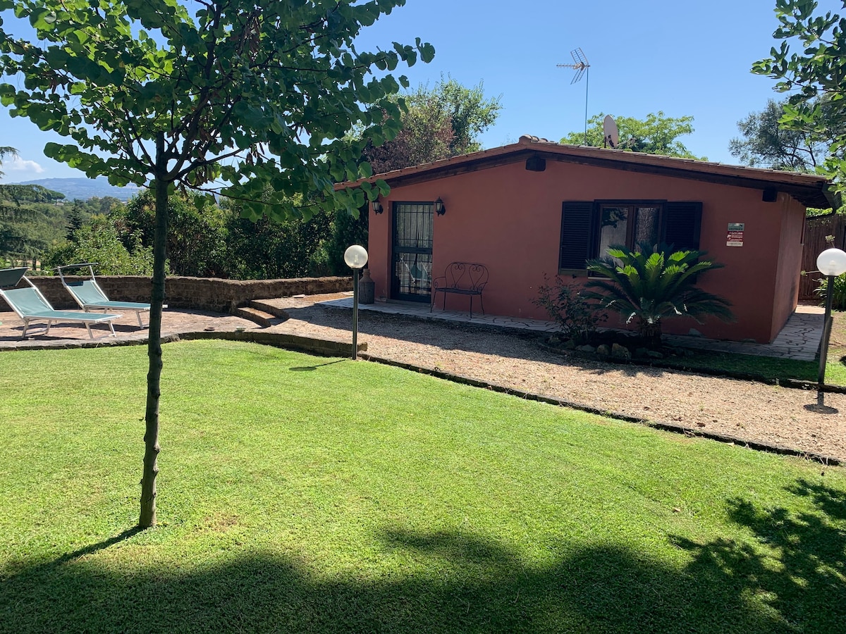 A charming depandance is visible, nestled amid lush greenery. The exterior features a terracotta facade, complemented by large windows. A well-maintained garden surrounds the space, with lounge chairs positioned near a refreshing pool, creating an inviting area for relaxation.