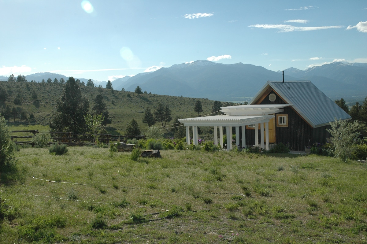 A charming cottage is set amidst a spacious grassy area, with mountain ranges visible in the background. The structure features a porch supported by white columns, enhancing the rural landscape and providing an inviting transition to the outdoors.