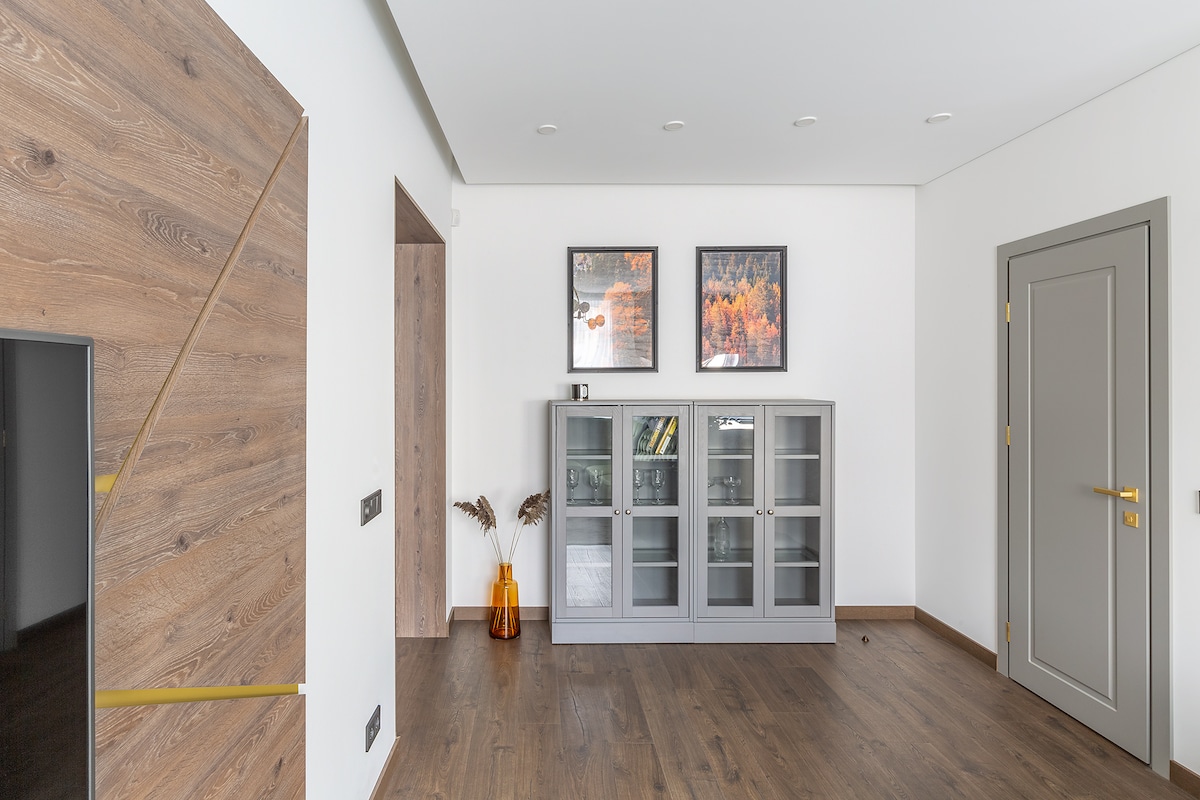 A spacious living room features light wood flooring and a neutral color palette. Two framed artworks are displayed on the wall above a gray cabinet with glass doors. A decorative vase with dried flowers adds a touch of warmth to the room.