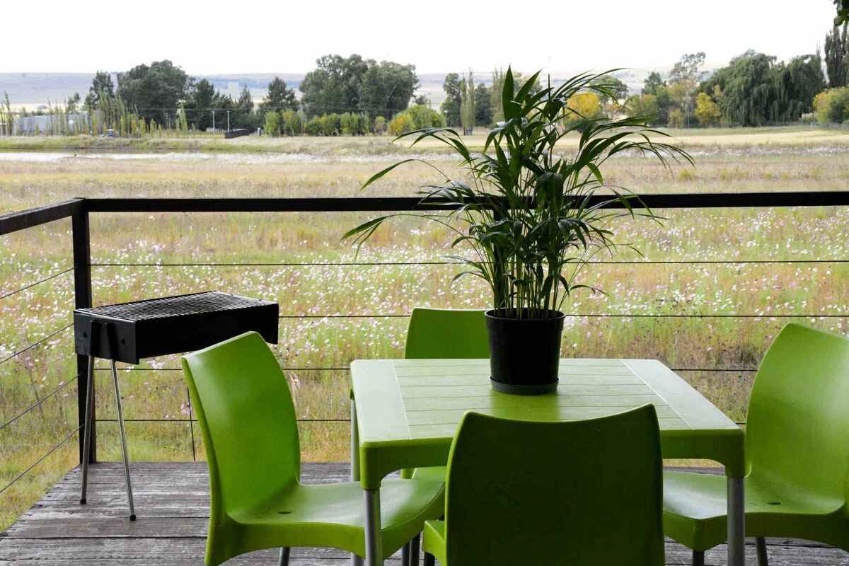 An outdoor seating area features a green table surrounded by four matching chairs. A potted plant sits at the center of the table. In the background, a scenic view of a grassy field is visible, complemented by trees lining the horizon.