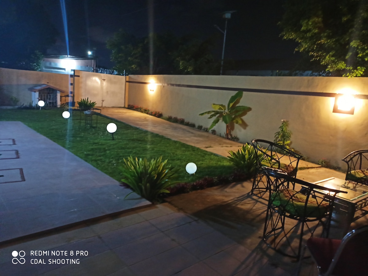 A well-maintained outdoor space is illuminated softly by garden lights in the evening. A small lawn features decorative plants, while a seating area with tables and chairs is positioned nearby, offering a relaxing spot in the serene environment.