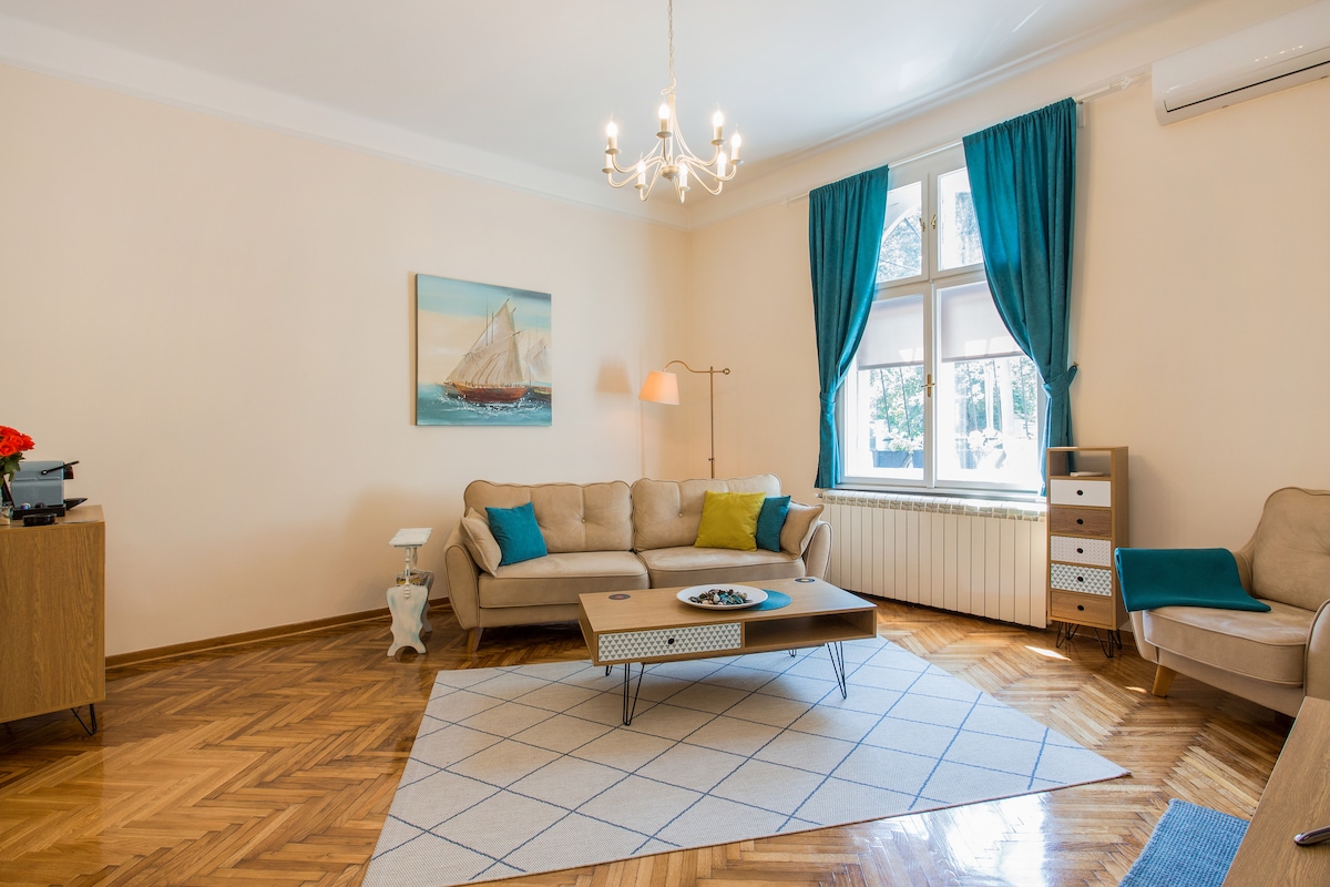 A spacious living area features a light-colored sofa adorned with accent pillows. A central coffee table sits atop a geometric rug, with a side table and a modern lamp nearby. Large windows allow natural light to fill the room, providing views of the outdoors.