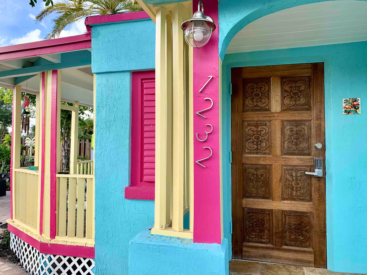 The bright blue exterior of Coastal Bungalow #1232 is complemented by vibrant yellow and pink accents. The entrance showcases a wooden door with intricate carvings and a unique door keypad for entry. Decorative elements such as the light fixture enhance the welcoming appearance.