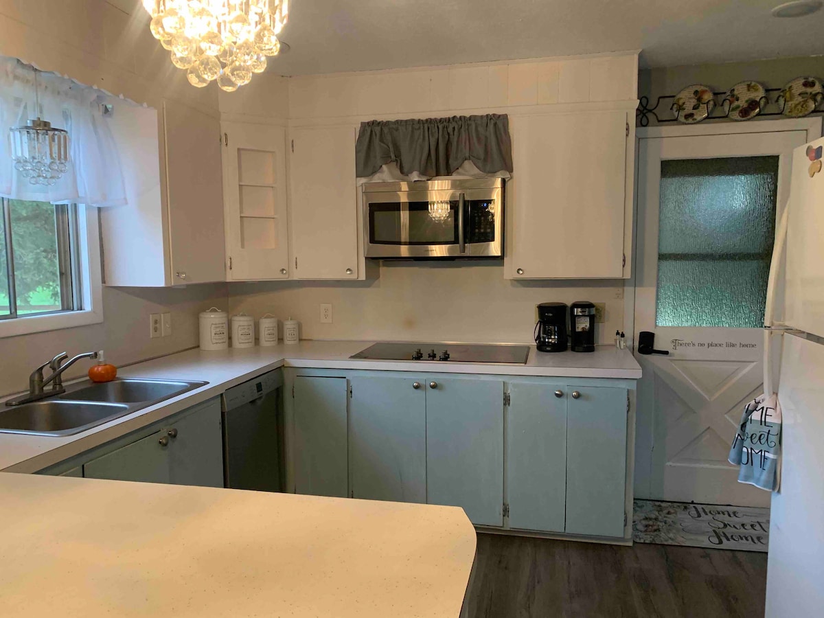 The kitchen features light-colored cabinetry and a spacious countertop. A modern microwave is mounted above the electric stove, and a coffee maker sits beside a double sink. Natural light enters through a window, complementing the room's bright and tidy appearance.