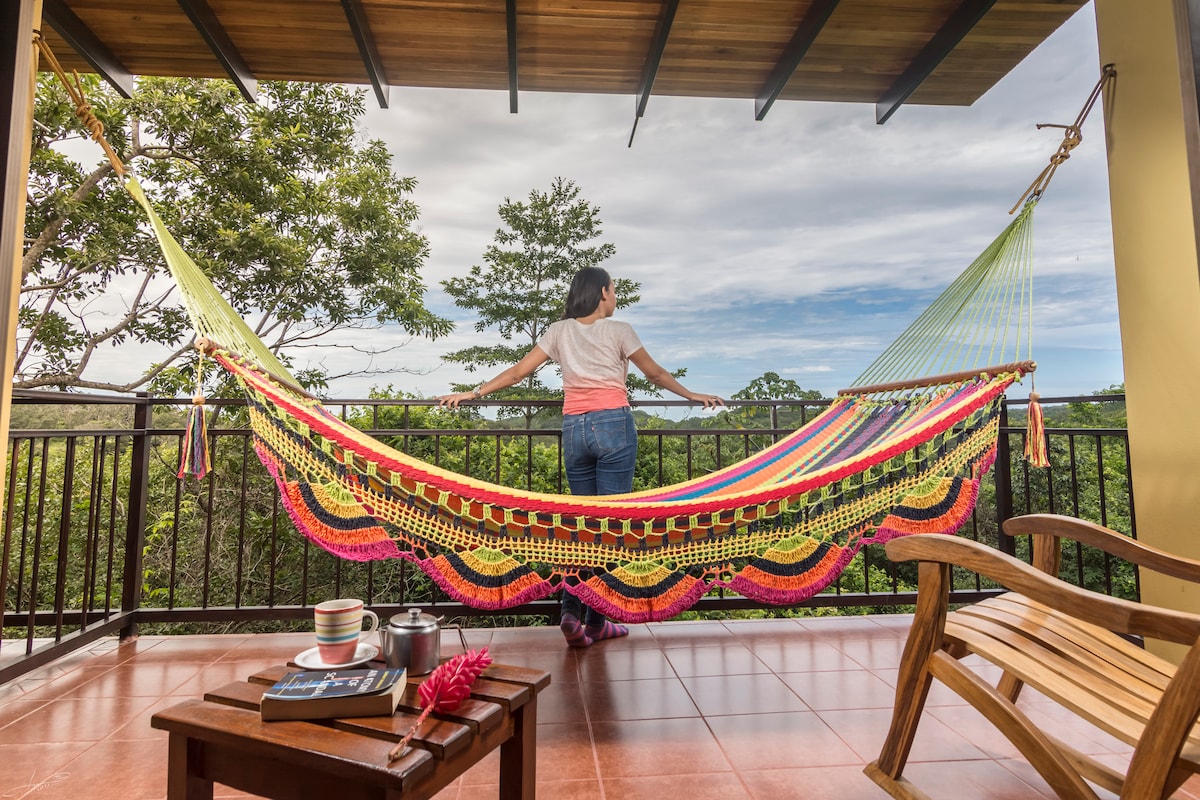 A colorful hammock is displayed on the balcony, overlooking lush greenery. A wooden rocking chair is positioned nearby, with a small table holding cups and a vibrant pink flower. A person stands relaxed in the hammock, enjoying the scenic mountain view.