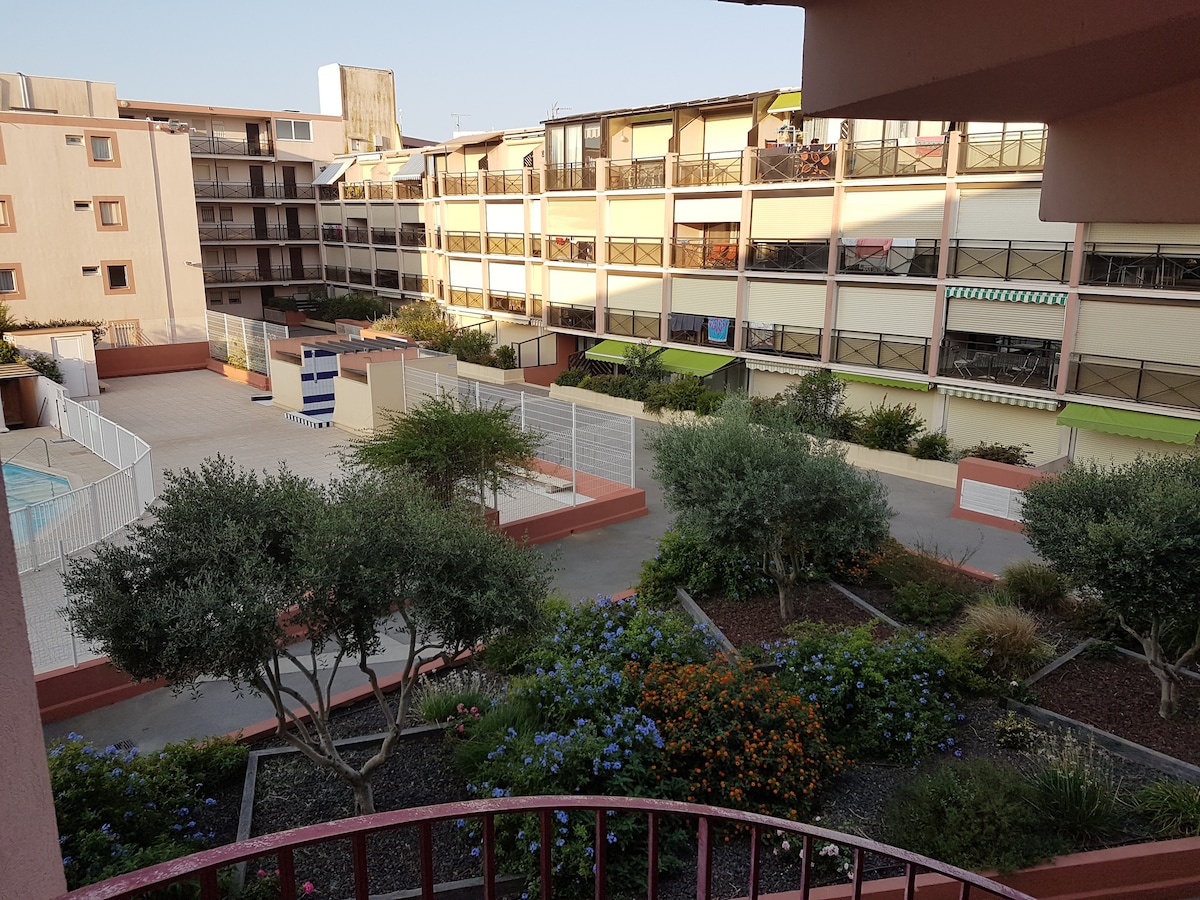A view from the terrace showcases a tranquil courtyard surrounded by well-maintained gardens filled with colorful flowers and greenery. The surrounding buildings feature multiple balconies and shaded awnings, creating a calm atmosphere. The area includes a swimming pool and is bordered by walking paths.