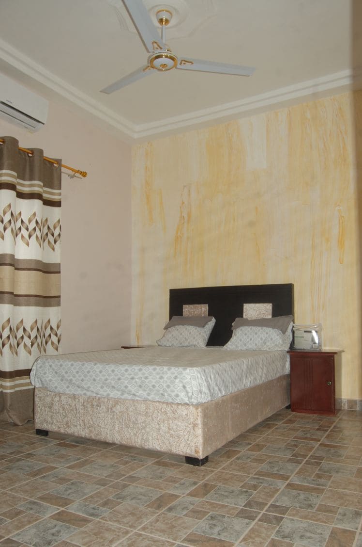 A sleeping area is furnished with a spacious bed featuring decorative headboards. Light-colored curtains with a patterned design frame a window, while a bedside table holds a lamp. The floor is covered with square tiles, complementing the soft tones of the walls.