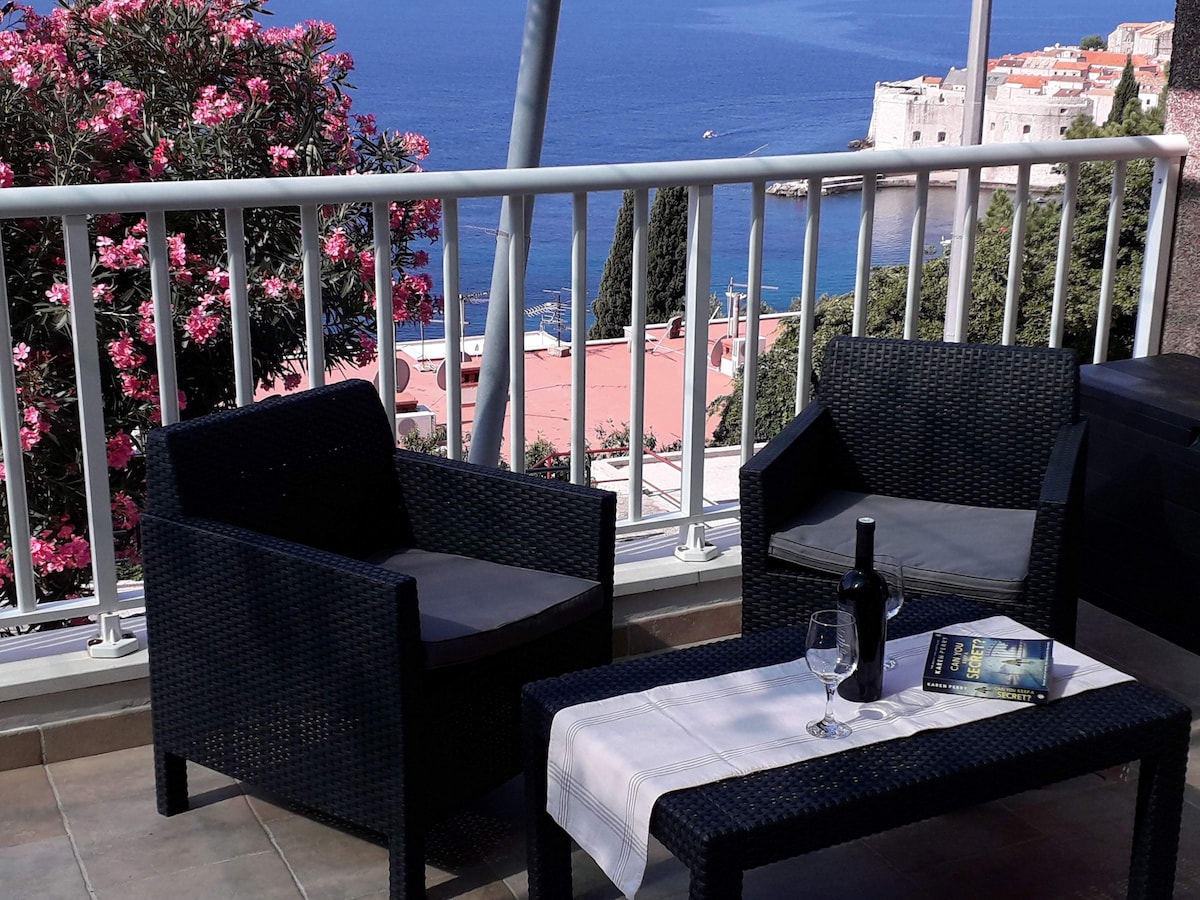 A terrace seating area features two woven chairs and a small table. A glass of wine and a book rest on the table, while a vibrant flowering plant can be seen nearby. The view includes the sea and part of Dubrovnik's historic architecture in the background.