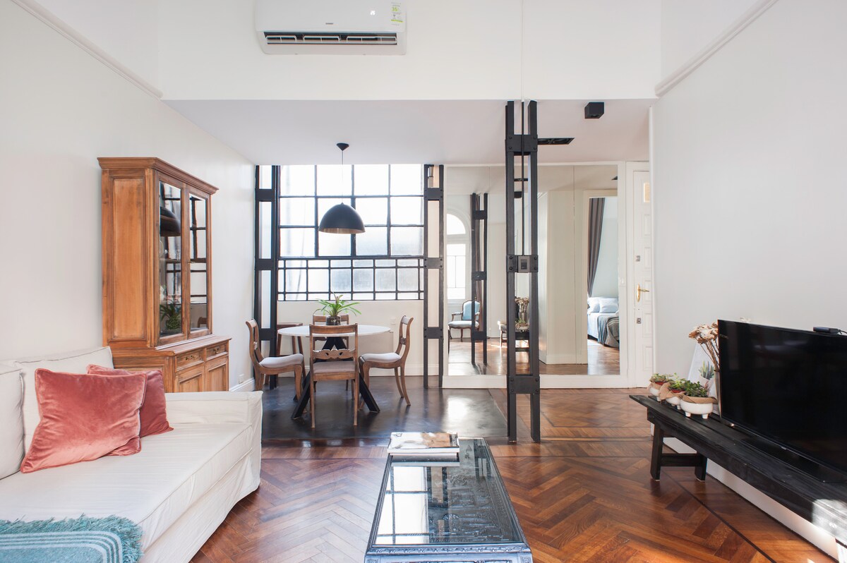 A bright living space features a comfortable sofa with decorative pillows and a wooden cabinet. A dining area with a round table and chairs is visible, and large windows allow natural light to illuminate the room. An elegant light fixture hangs above the dining table.
