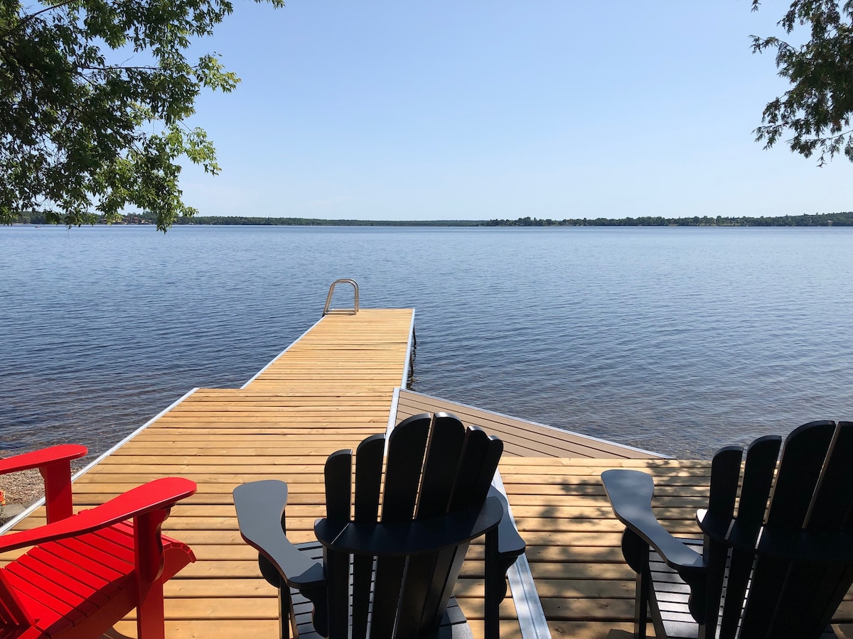 A wooden dock extends over calm water, providing access for swimming. Muskoka chairs in black and red are positioned on the lakefront deck, offering seating with unobstructed views of the serene landscape. Clear skies and distant trees frame the tranquil setting.