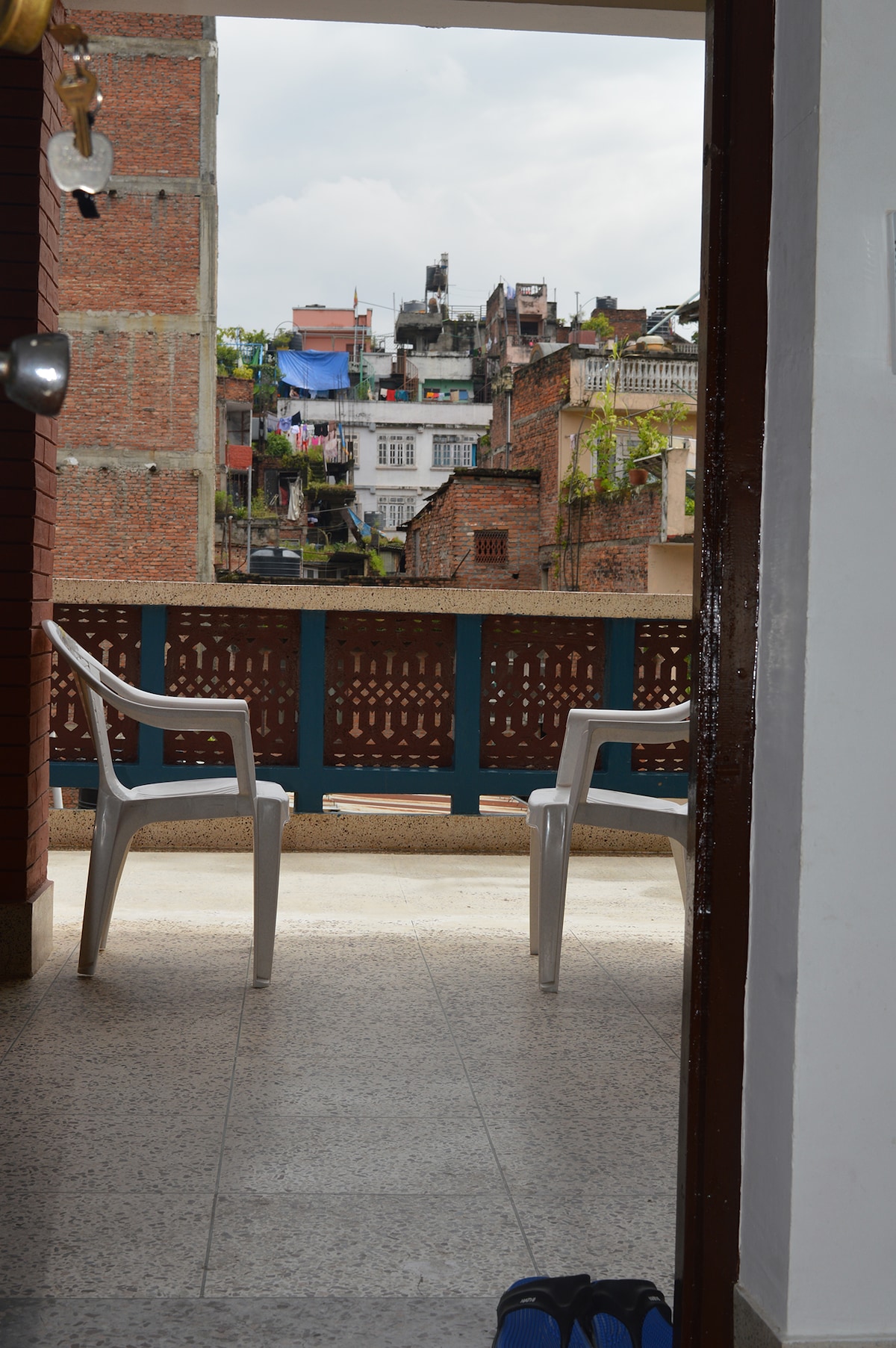 A sun terrace is visible, featuring two white plastic chairs positioned against a brick wall. The view includes neighboring buildings adorned with colorful laundry and green plants, creating a glimpse of the local environment.