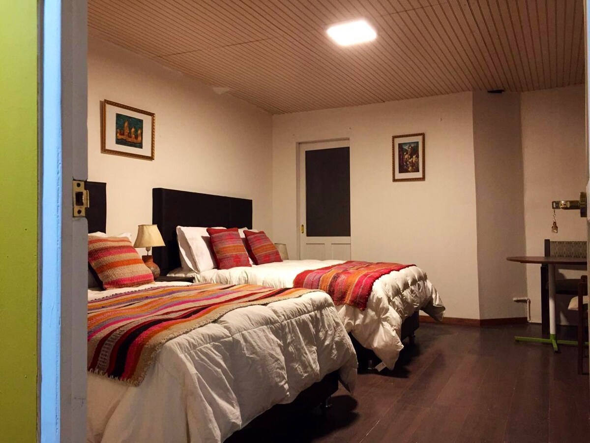 The spacious room features two comfortable double beds, each adorned with colorful woven blankets. A small bedside lamp provides soft lighting. A dining table is positioned at the far end, and a door leads to the balcony, enhancing the room's overall openness.