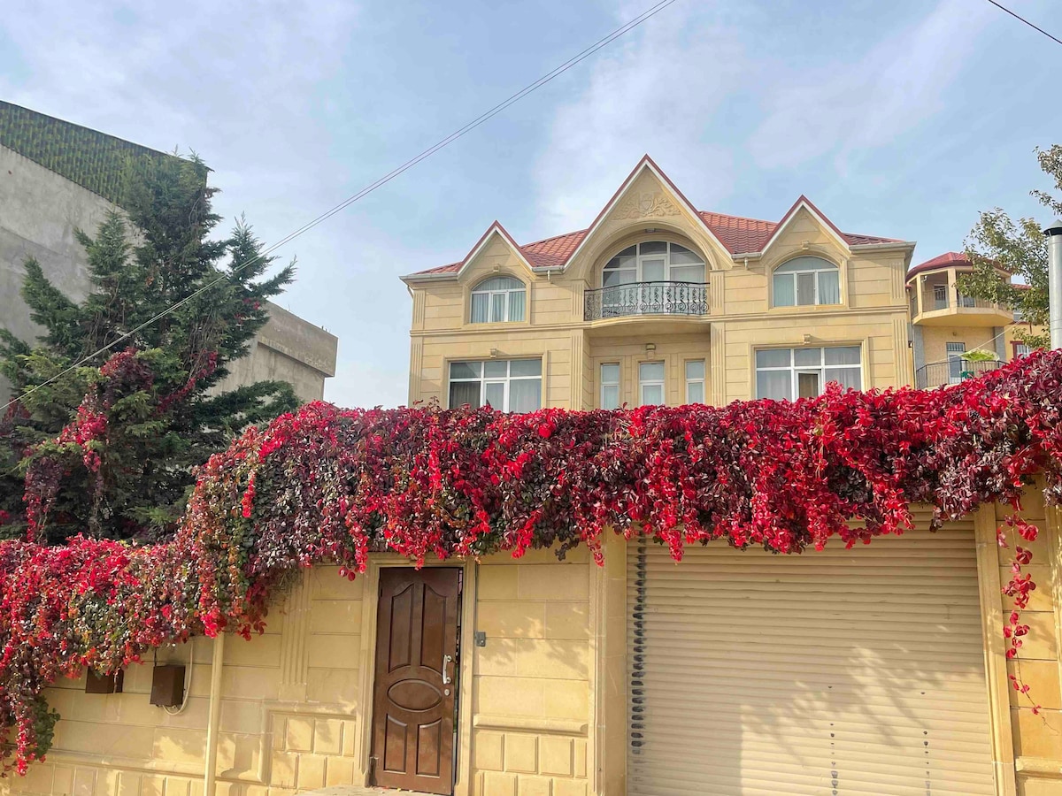A charming building is featured, showcasing a façade adorned with vibrant flowers cascading from the top. Large windows provide ample natural light, and a decorative roof peaks above the main entrance, which is framed by lush greenery.