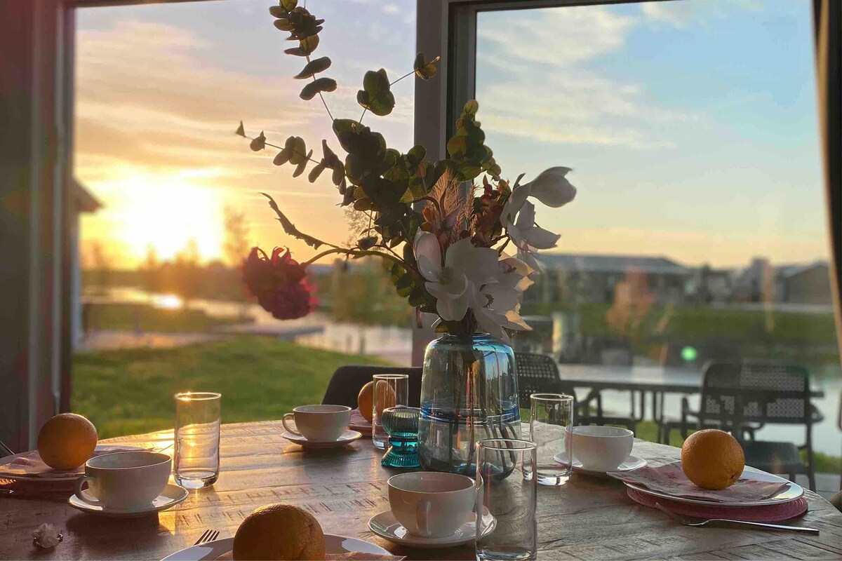 A dining area is set with a rustic wooden table featuring utensils, glasses, and bowls filled with fruit. A vibrant flower arrangement is centered on the table, while large windows provide a view of a sunset illuminating the serene waters outside.