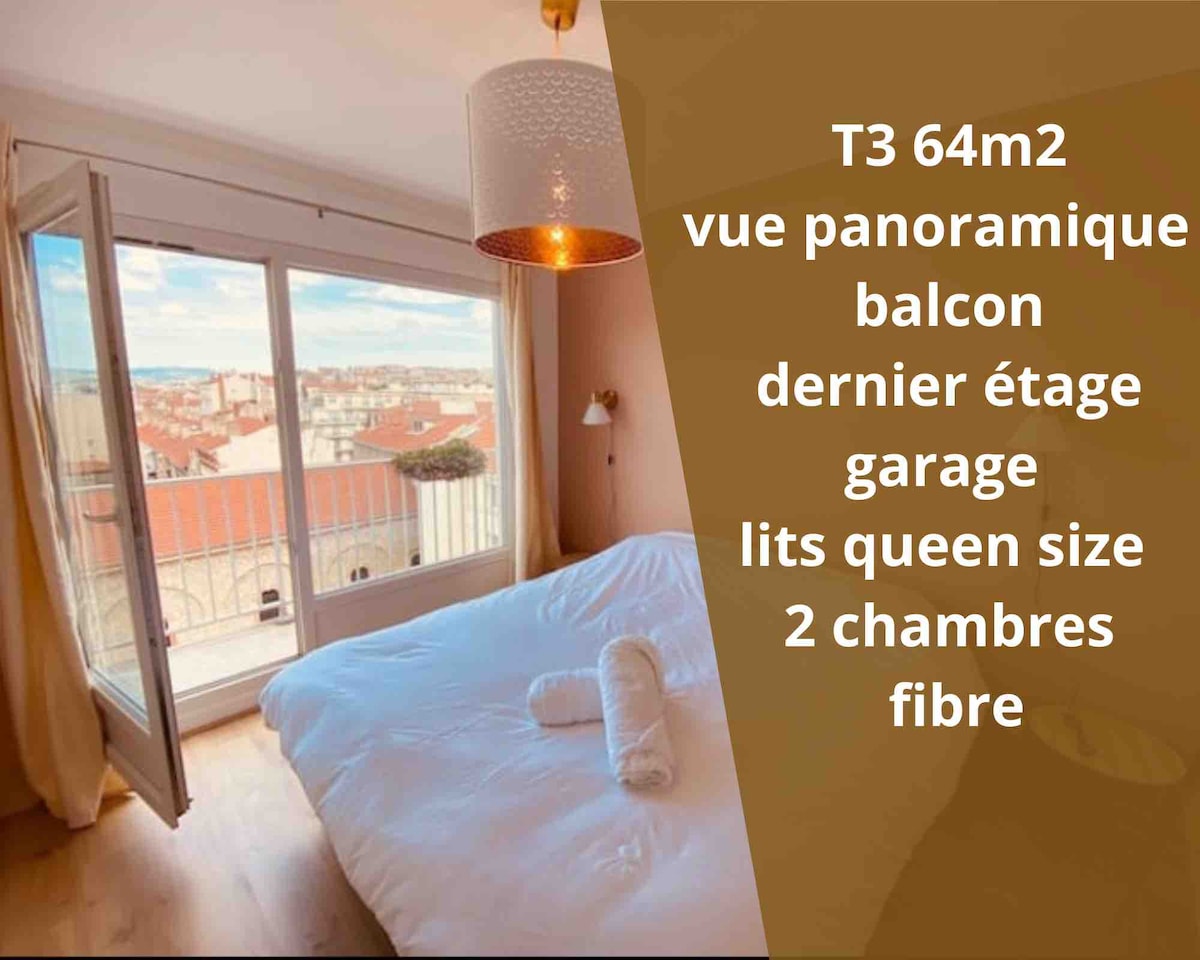 A bright bedroom is highlighted by large glass doors leading to a balcony, offering panoramic views of the rooftops. A spacious bed is centered in the room, with warm wooden flooring and simple décor enhancing the calm atmosphere.