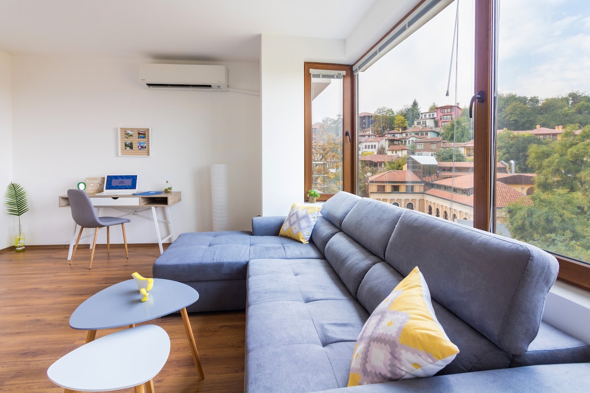 A spacious living area features a large gray sectional sofa accented with decorative pillows. A modern desk with a computer is positioned beside a window, offering views of the historical buildings outside. Natural light floods the room, complemented by warm wood flooring.