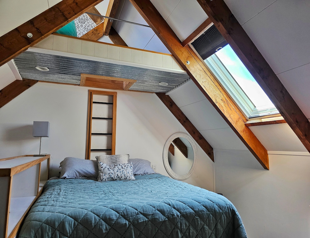 A cozy sleeping area features a spacious bed covered in a textured teal quilt. Natural light floods the room through overhead skylights, highlighting the wooden ceiling beams. A round mirror and an accent lamp add functional elements to the inviting space.
