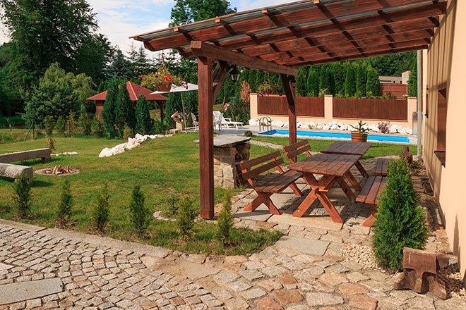 A shaded outdoor seating area is featured beneath a wooden pergola, showcasing a sturdy picnic table with attached benches. The well-maintained lawn surrounds the space, with neatly arranged stone pathways leading towards a swimming pool visible in the background.