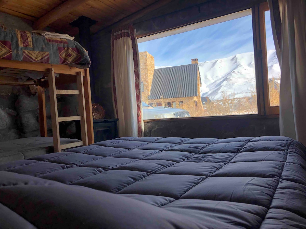A cozy sleeping area includes a spacious bed covered with a dark comforter. A large window offers a view of the snow-capped mountains outside, while sunlight fills the room. A wooden bunk bed stands in the corner, complemented by rustic decor.