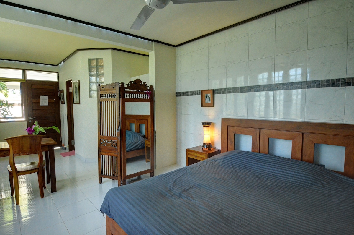 A spacious bedroom is furnished with a large bed and wooden decor. A dining table sits nearby, complemented by a traditional Indonesian room divider. Natural light enters through large windows, with a fan overhead providing ventilation.