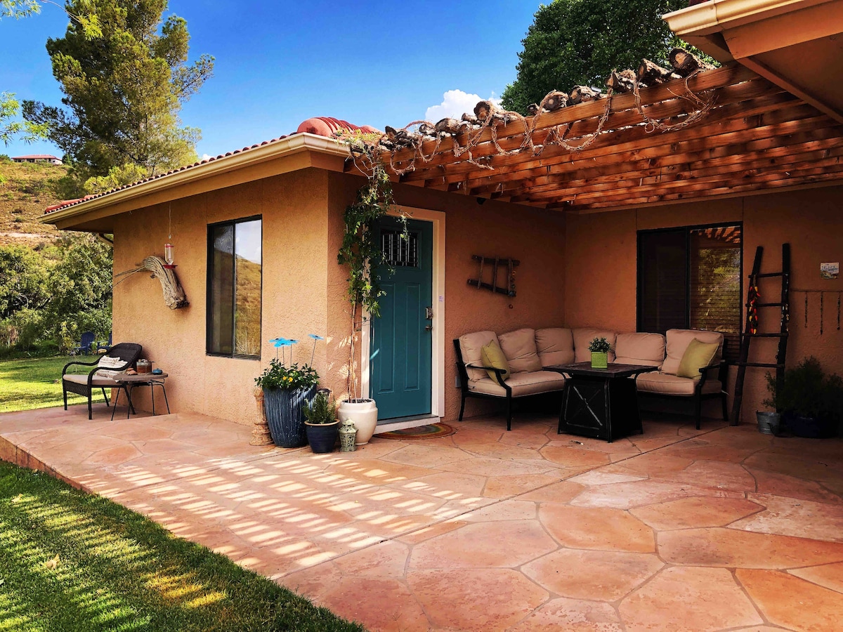 A charming patio area is furnished with a cozy couch and coffee table, shaded by a pergola. Lush greenery surrounds the space, accented by potted plants. The entrance features a turquoise door, leading into the guest wing, while geometric patterns cast by sunlight are visible on the stone floor.