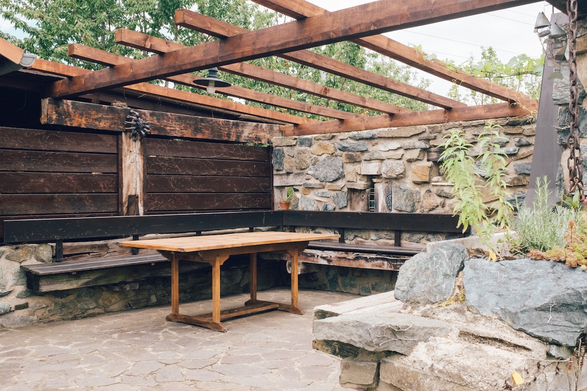An outdoor seating area is framed by rustic wooden beams and stone walls. A wooden table is positioned in the center, surrounded by built-in benches. Green plants are visible among the natural stone elements, creating a serene space for gatherings.