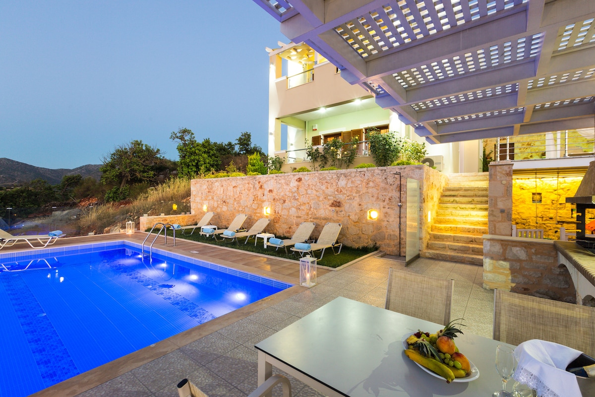 The outdoor space features a private heated pool surrounded by sun loungers, with a dining table set for al fresco meals. Soft lighting enhances the stone walls and staircase leading to the villa, with lush greenery in the background under a twilight sky.