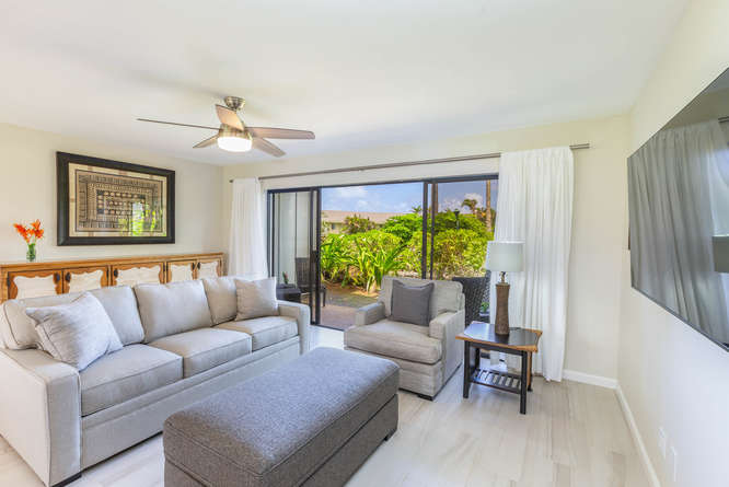 A comfortable living room is featured with a light grey sectional sofa and an accent chair. Large glass doors lead to a private outdoor space surrounded by greenery, while a fan overhead adds a functional touch. A neutral coffee table complements the decor.