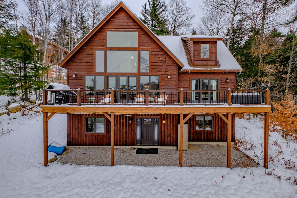 The exterior showcases a wooden cabin-style home with large windows that capture the surrounding landscape. A deck spans the front, supported by wooden pillars, with accessible entry to the main door. Snow blankets the ground, indicating a serene winter setting.