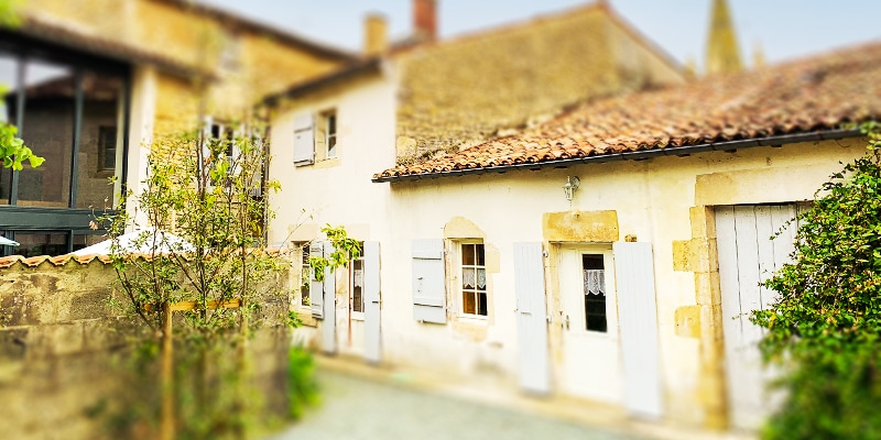 A charming exterior view of the house displays a traditional stone façade with beige tones and white shutters. The roof is covered with terracotta tiles. A small garden area is visible in front, featuring green foliage that adds a touch of nature.