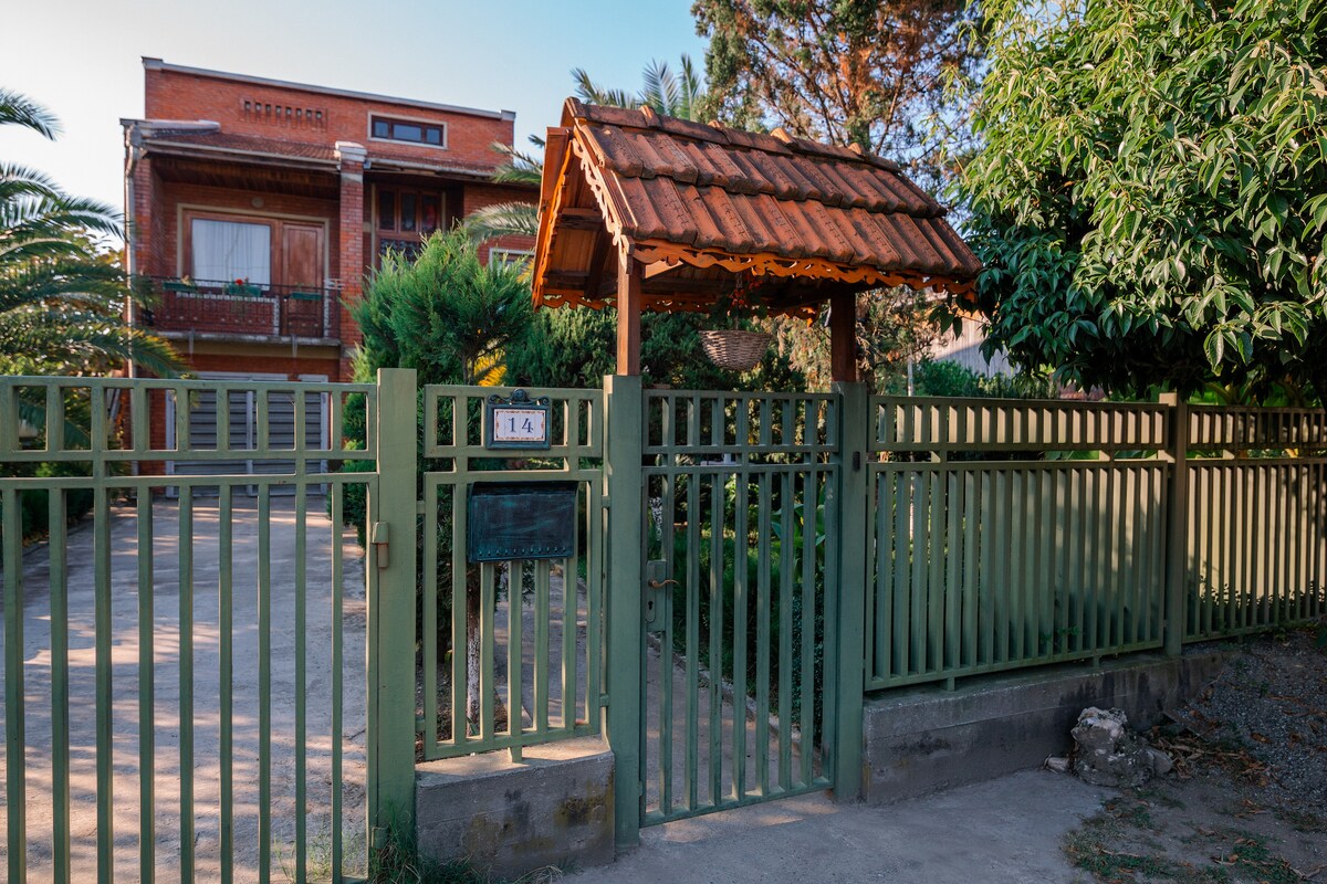 A green gate opens to a gravel path leading to a multi-story brick house surrounded by trees and shrubs. A wooden awning with a red roof provides shelter over the entryway. The tranquil setting suggests a private and welcoming ambiance.