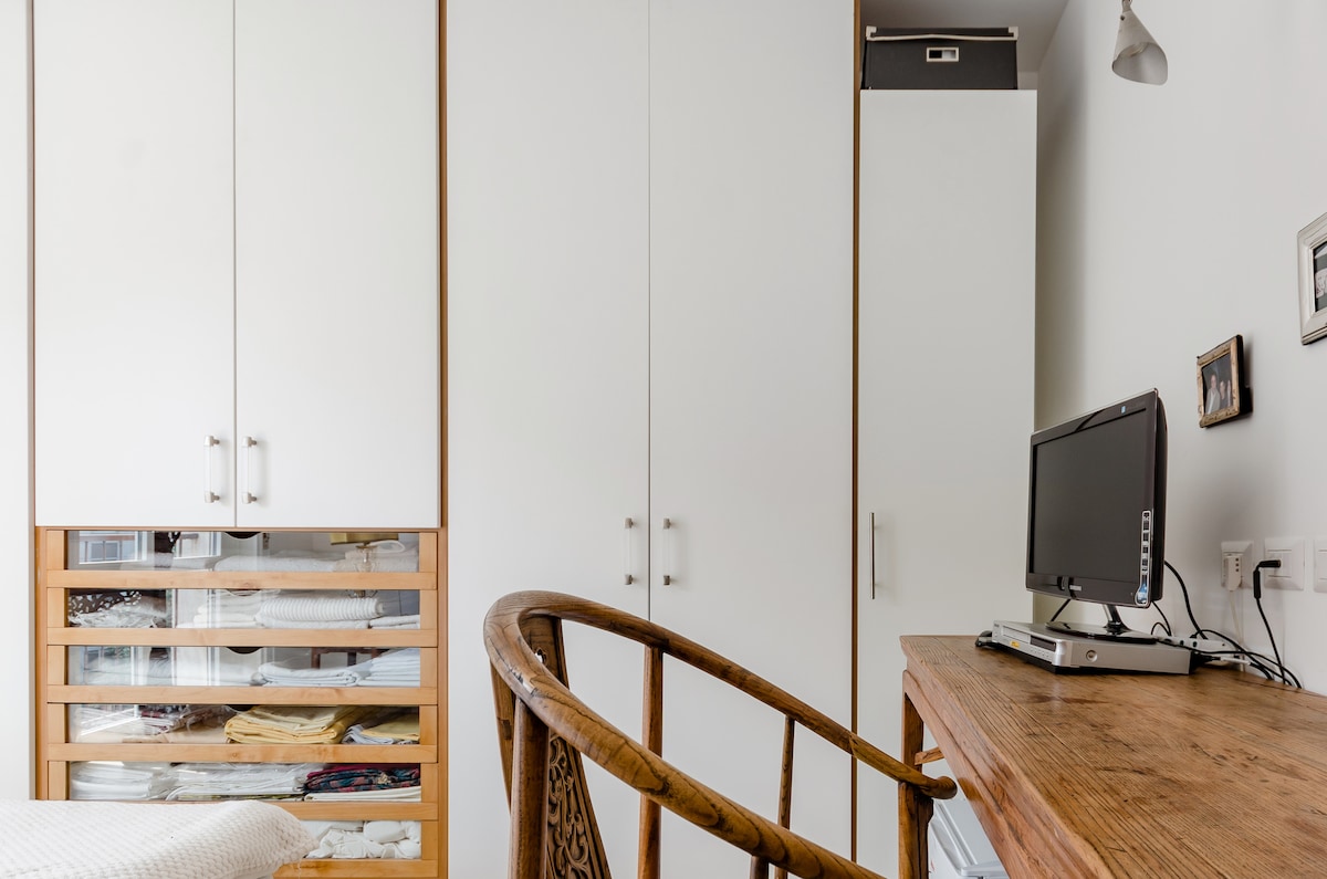 A bright and organized sleeping area is presented, featuring built-in wardrobes with wooden accents. A wooden desk is accompanied by a chair, while a computer monitor sits on the desk. Natural light illuminates the space, creating a functional yet cozy atmosphere.