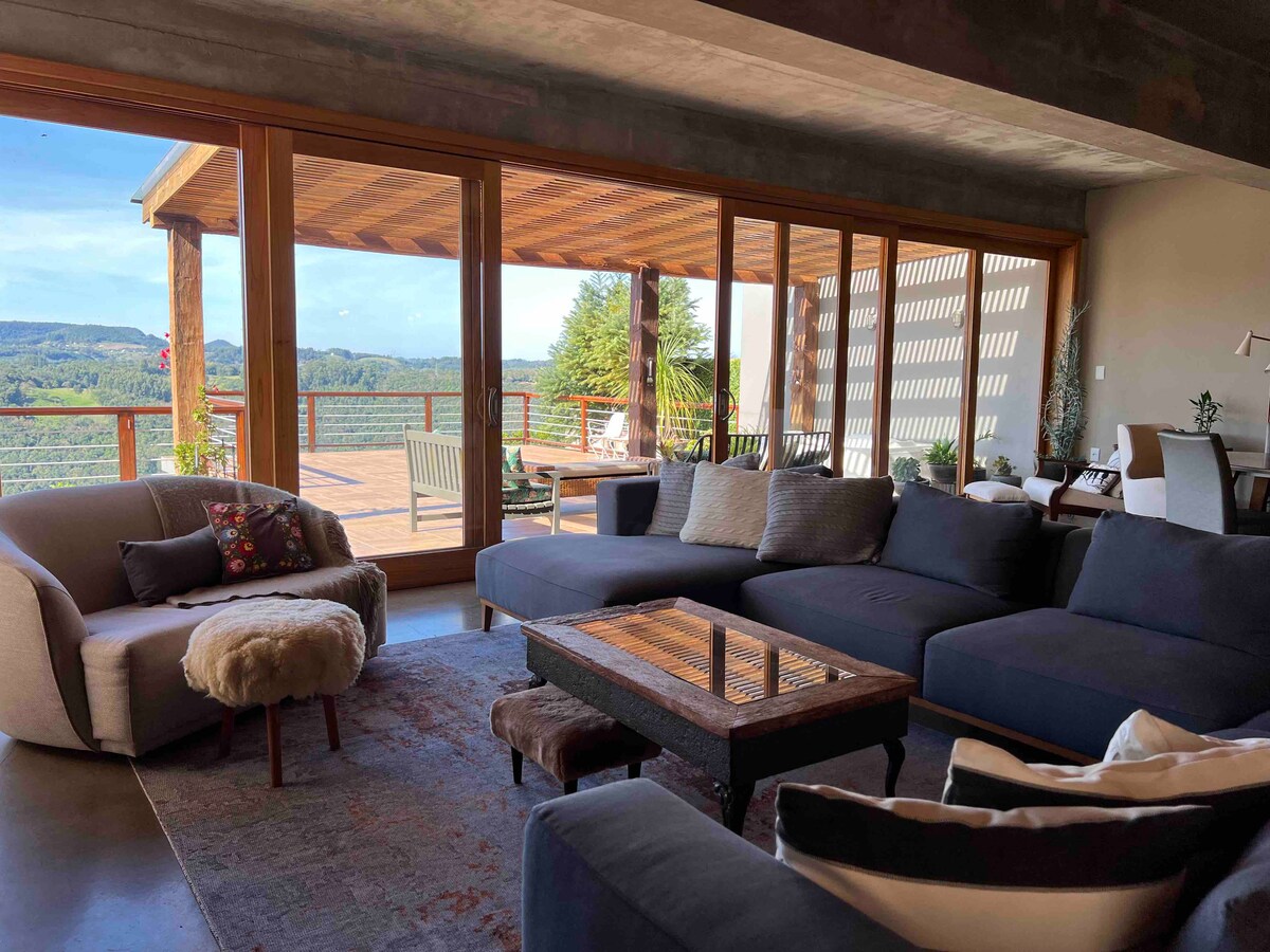 A spacious living area is furnished with a large, sectional sofa and a cozy accent chair. Expansive glass doors provide an unobstructed view of the outdoor terrace with seating, surrounded by lush greenery and distant hills under a clear sky.