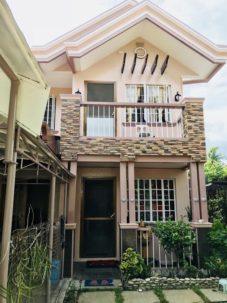 The exterior of a two-story house features a combination of light and stone-colored walls. Windows with white grilles provide views into the interior. A well-maintained garden with small plants and decorative stones lines the pathway leading to the entrance.