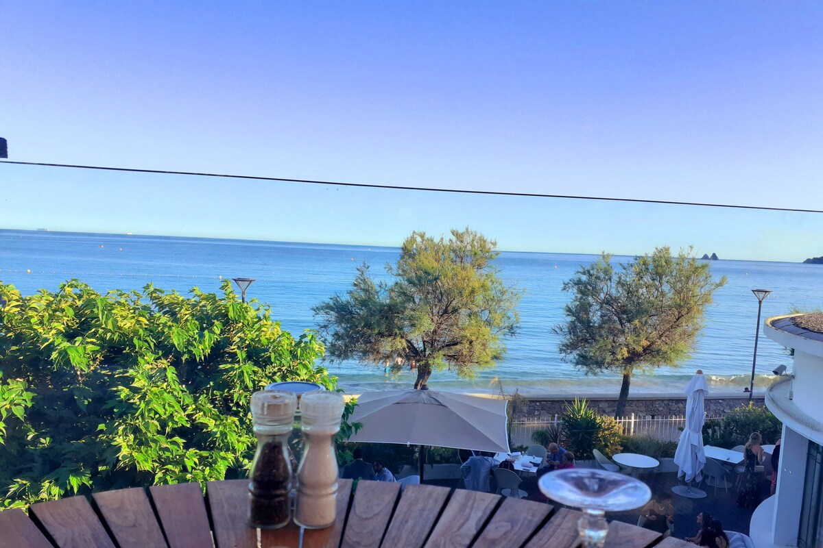 The image features a serene view of the sea, visible through a glass railing. In the foreground, condiments are neatly arranged on a wooden table, while the beach and trees line the shore, complementing the calm water and clear skies.