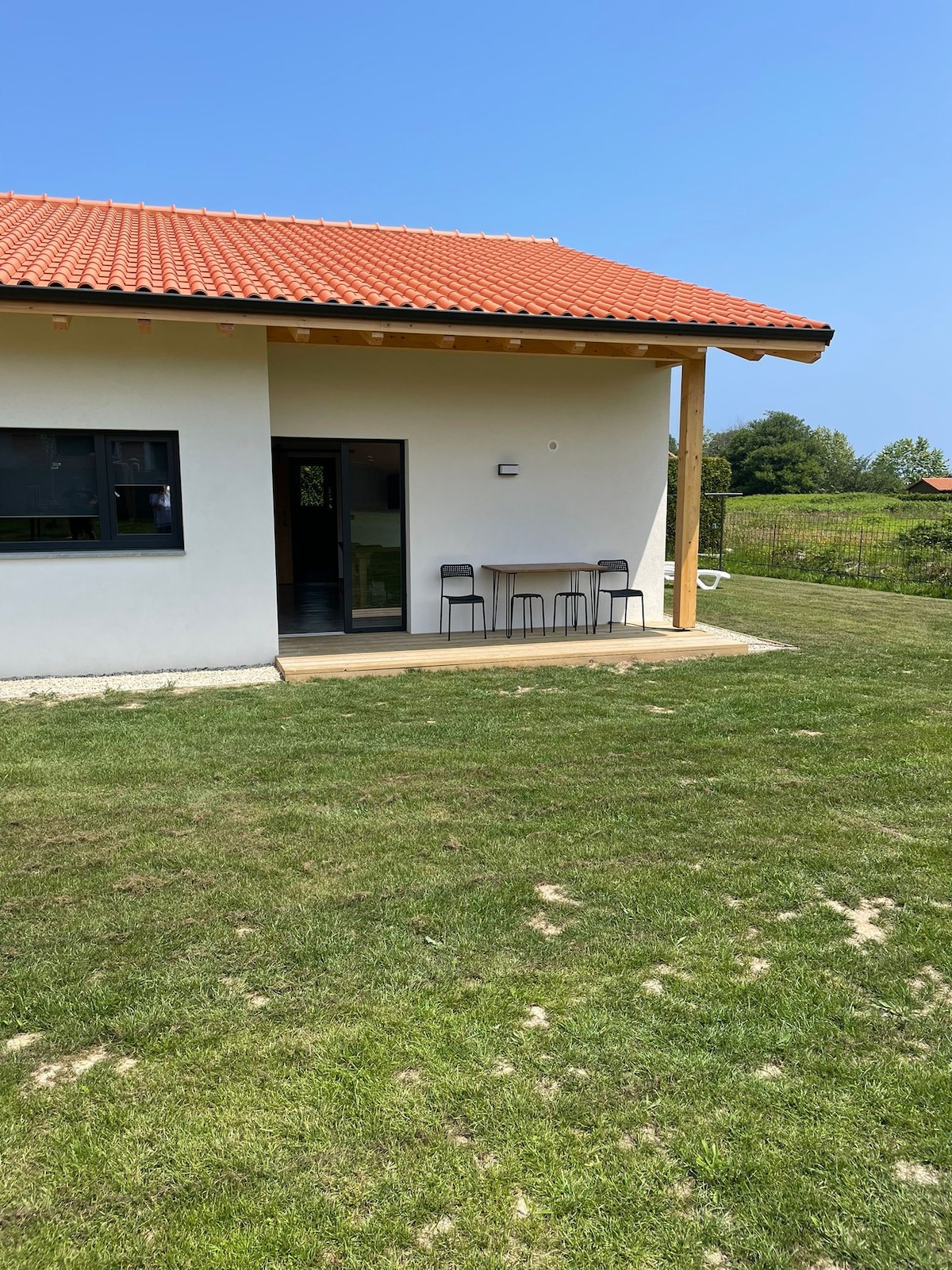 A modern building is depicted with a red-tiled roof and a white exterior. A small outdoor patio features a table and four chairs, set against a backdrop of well-maintained lawn and greenery. Clear skies and sunlight enhance the serene environment.