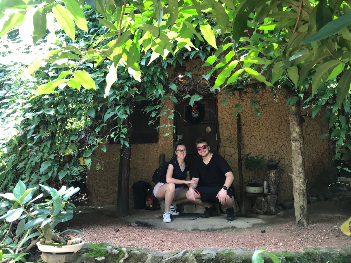 A charming earthen cottage is partially hidden by lush greenery, featuring a thatched roof and walls adorned with vines. Guests are seated on a rustic bench outside the entrance, surrounded by natural textures and a serene atmosphere.