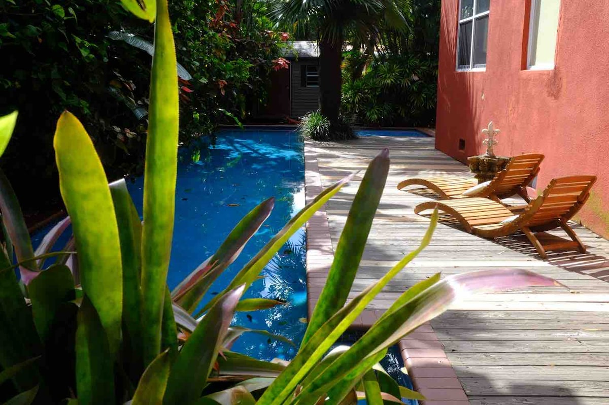 A serene pool area is framed by lush tropical foliage, showcasing a well-maintained pool alongside wooden deck space. Two lounge chairs are positioned beside the water, inviting relaxation amidst the greenery.