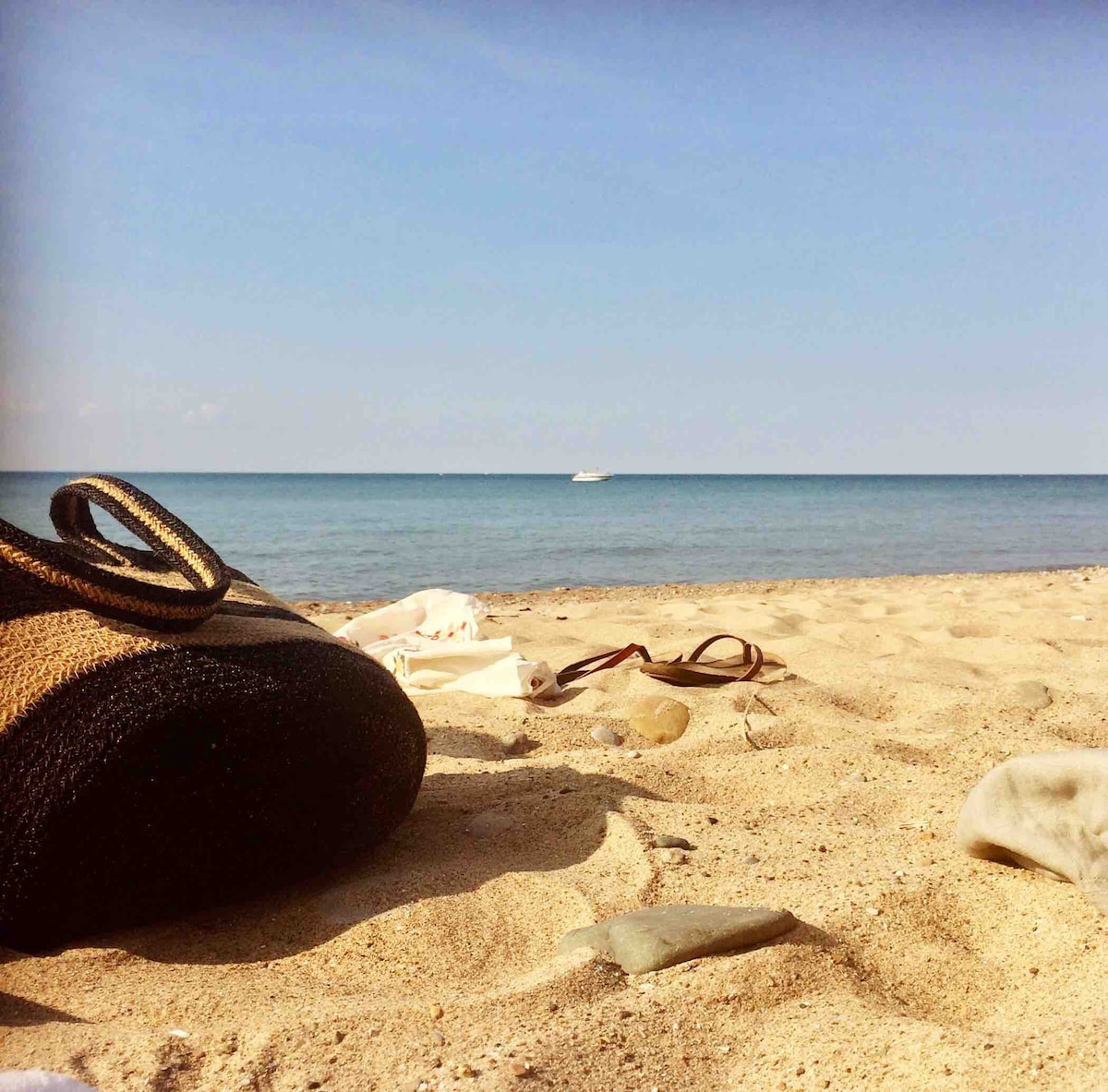 A sandy beach is captured under a clear blue sky, with gentle waves lapping at the shoreline. A woven bag lies on the sand, surrounded by scattered light-colored fabric and smooth stones, creating a serene and relaxed coastal atmosphere.
